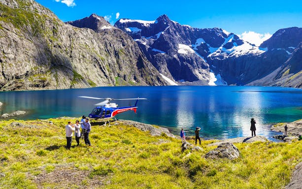 Helicopter landing by a lake in Milford Sound with people exploring the scenic mountain landscape.