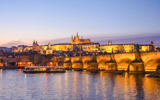 Prague Castle and Charles Bridge illuminated at dusk, reflecting on the Vltava River.
