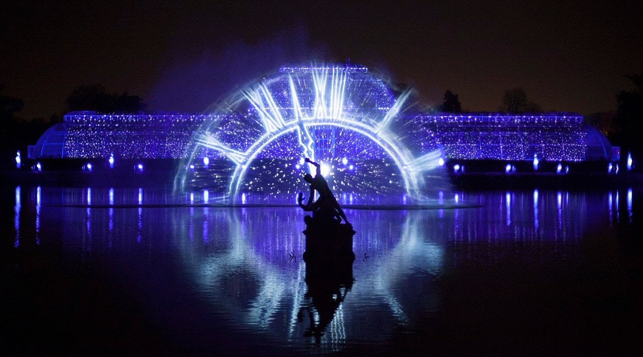 Light display at Kew Gardens with illuminated glasshouse and water reflection.