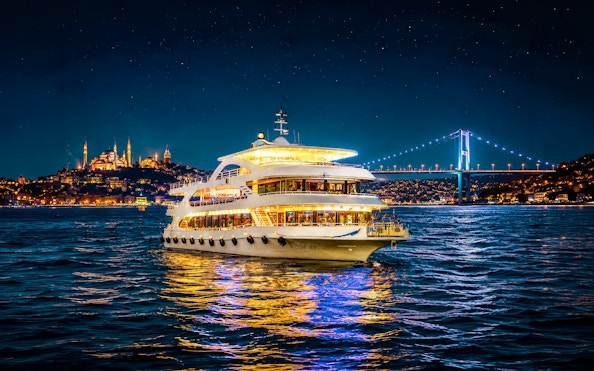 Cruise ship on Bosphorus strait at night with illuminated bridge and cityscape.