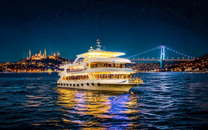 Cruise ship on Bosphorus strait at night with illuminated bridge and cityscape.