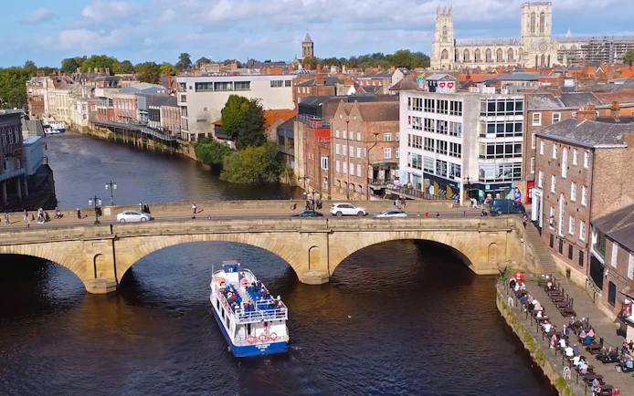 York sightseeing cruise passing under a historic stone bridge with York Minster in the background.