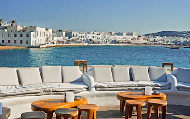 Outdoor seating overlooking Mykonos waterfront with white buildings and blue sea.