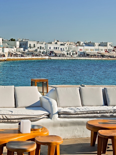 Outdoor seating overlooking Mykonos waterfront with white buildings and blue sea.