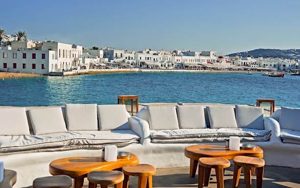 Outdoor seating overlooking Mykonos waterfront with white buildings and blue sea.