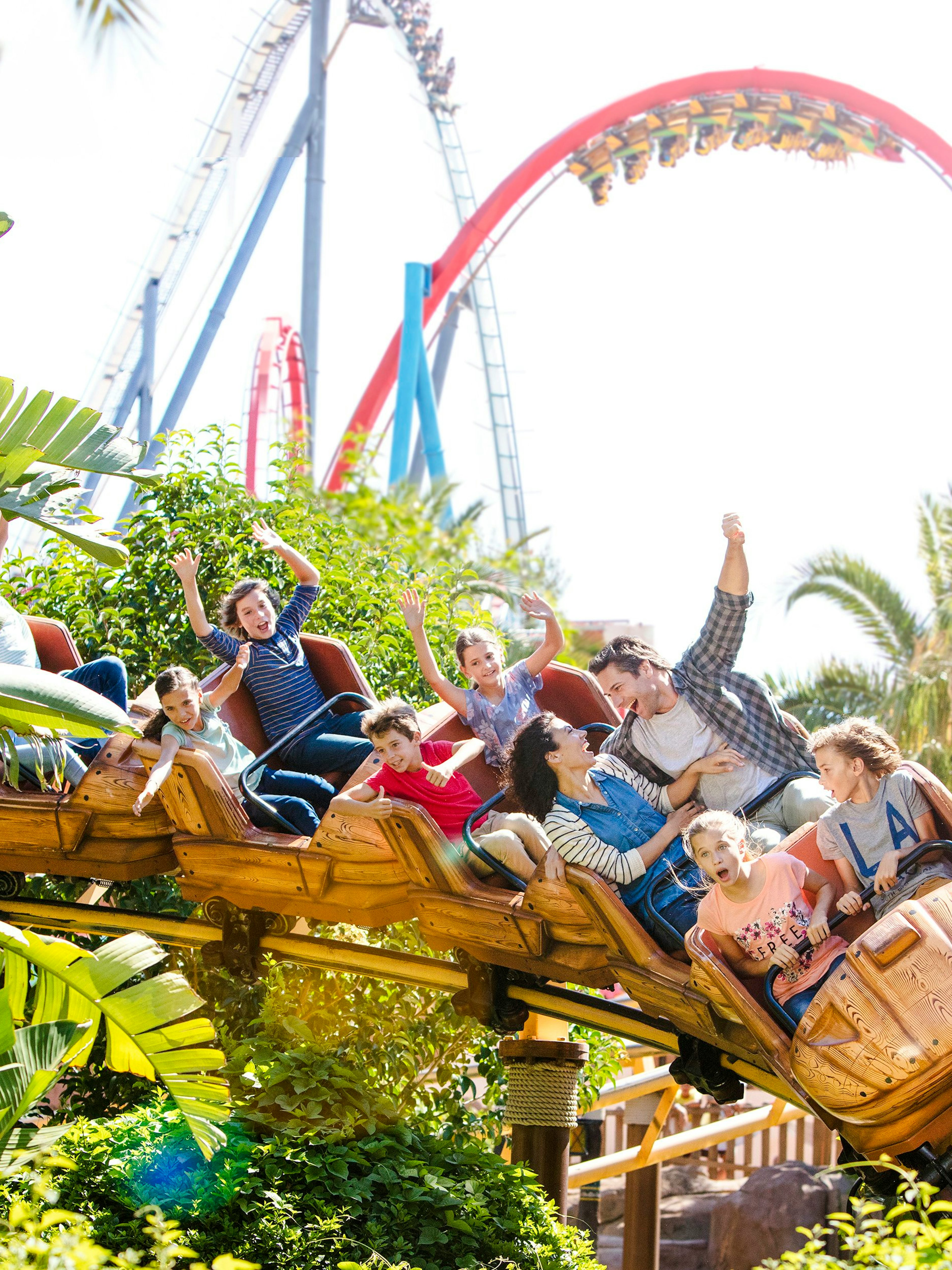 Families enjoying the Tami Ride at Portaventura amidst lush greenery and roller coaster loops.