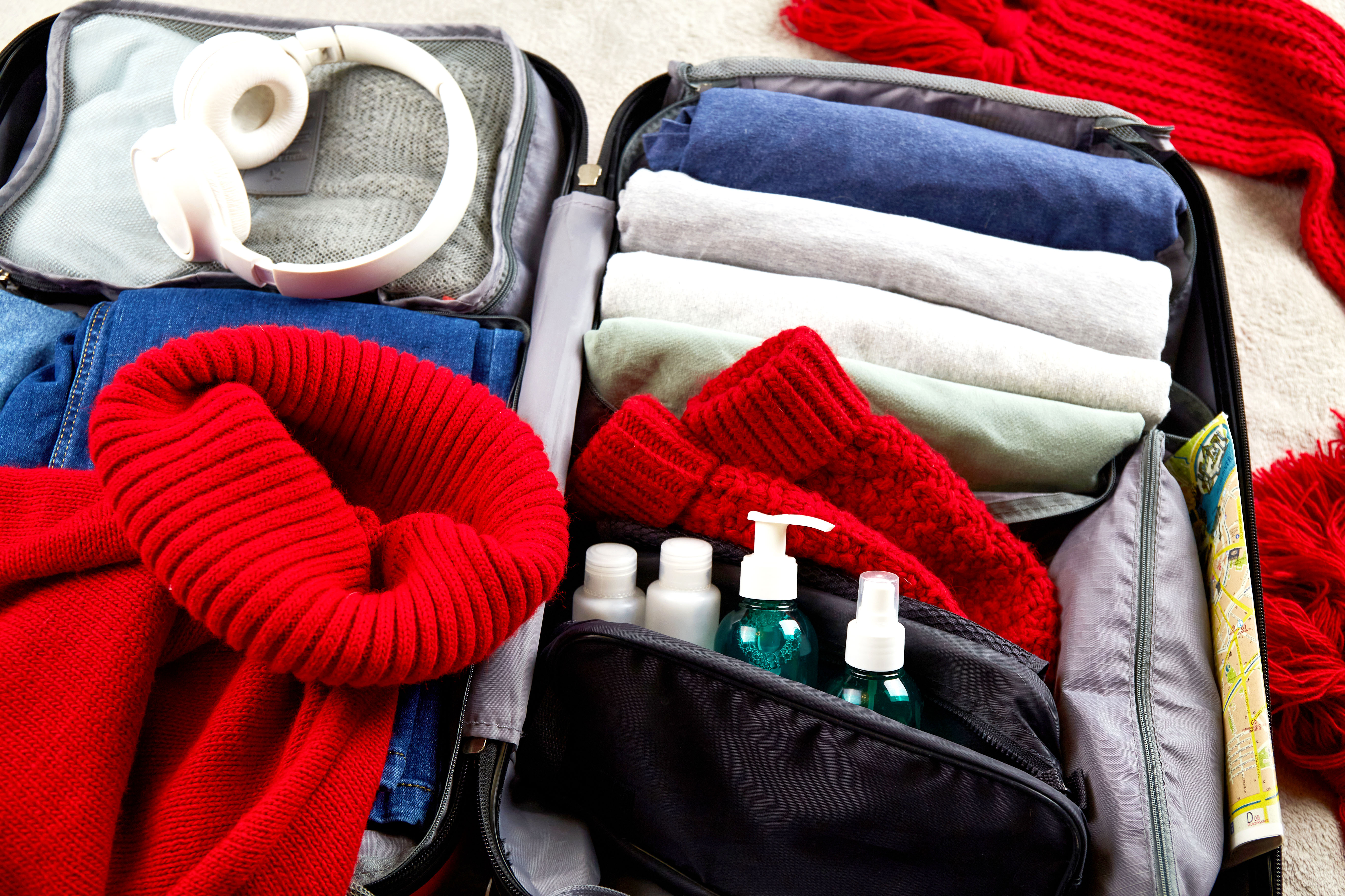 Open suitcase with winter clothes, red scarf, headphones, and toiletries for winter travel packing.