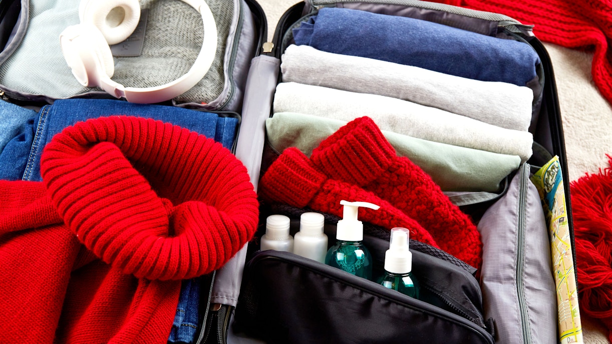 Open suitcase with winter clothes, red scarf, headphones, and toiletries for winter travel packing.