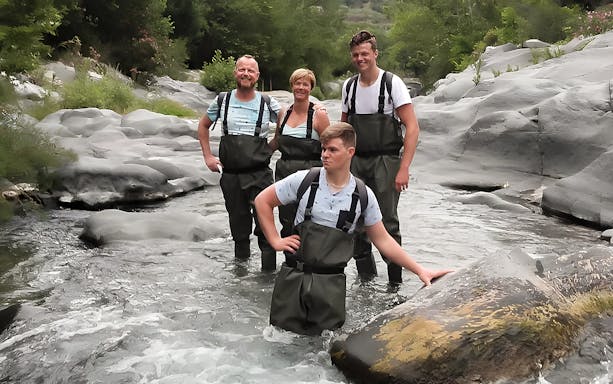 Group river trekking in Gole Alcantara, Sicily, wearing waders amidst rocky stream.