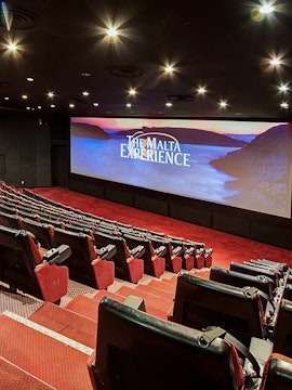 Interior of The Malta Experience theater with empty red seats and large screen displaying the title.
