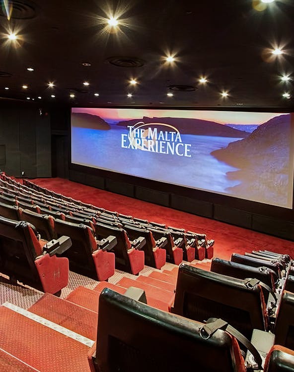 Interior of The Malta Experience theater with empty red seats and large screen displaying the title.