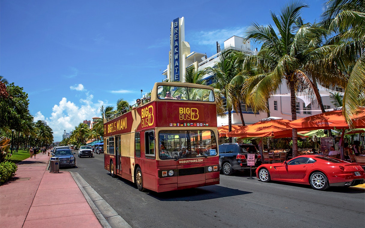 Open-top double-decker bus touring Miami's Ocean Drive with palm trees and art deco buildings.