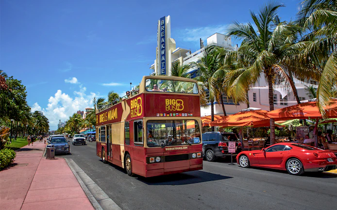 Open-top double-decker bus touring Miami's Ocean Drive with palm trees and art deco buildings.