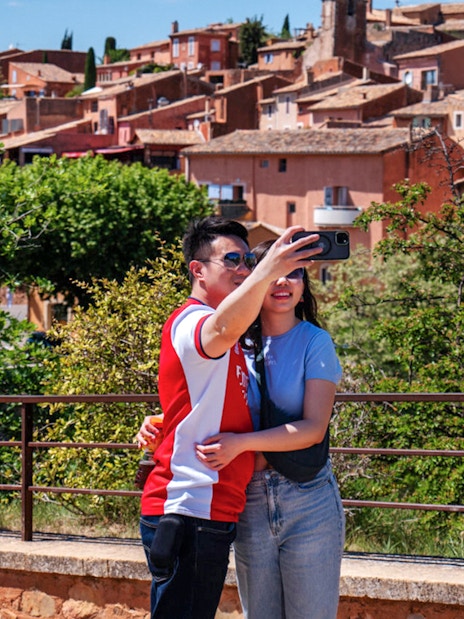 Couple taking a selfie with the colorful village of Roussillon, Provence in the background.
