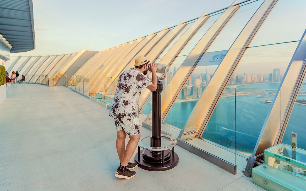 Man using binoculars at The View At The Palm, overlooking Dubai skyline and coastline.