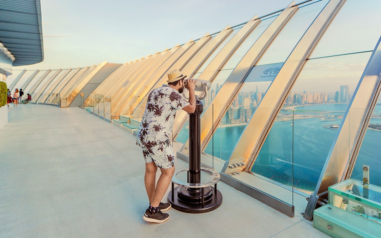 Man using binoculars at The View At The Palm, overlooking Dubai skyline and coastline.