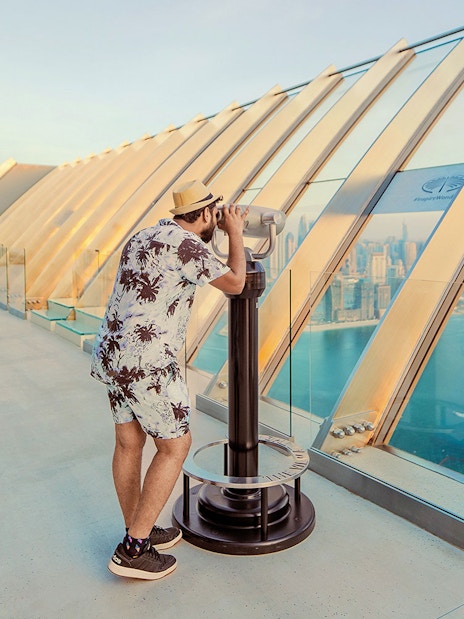 Man using binoculars at The View At The Palm, overlooking Dubai skyline and coastline.