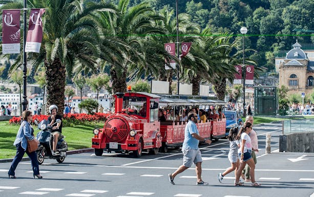 Red tourist train on a street in San Sebastian with palm trees and pedestrians nearby.