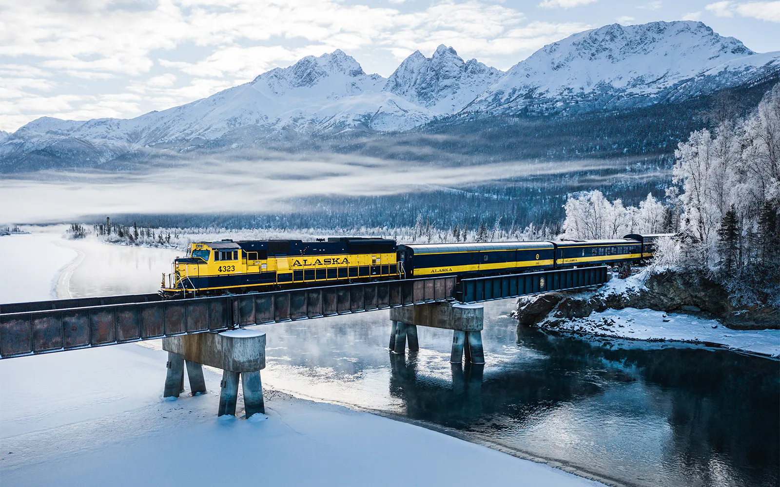 Aurora Winter Train crossing snowy Alaskan landscape with mountains in the background.