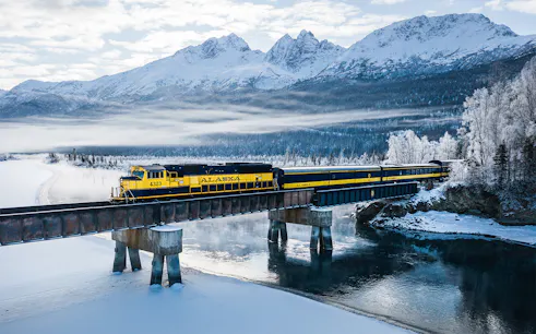 Aurora Winter Train crossing snowy Alaskan landscape with mountains in the background.