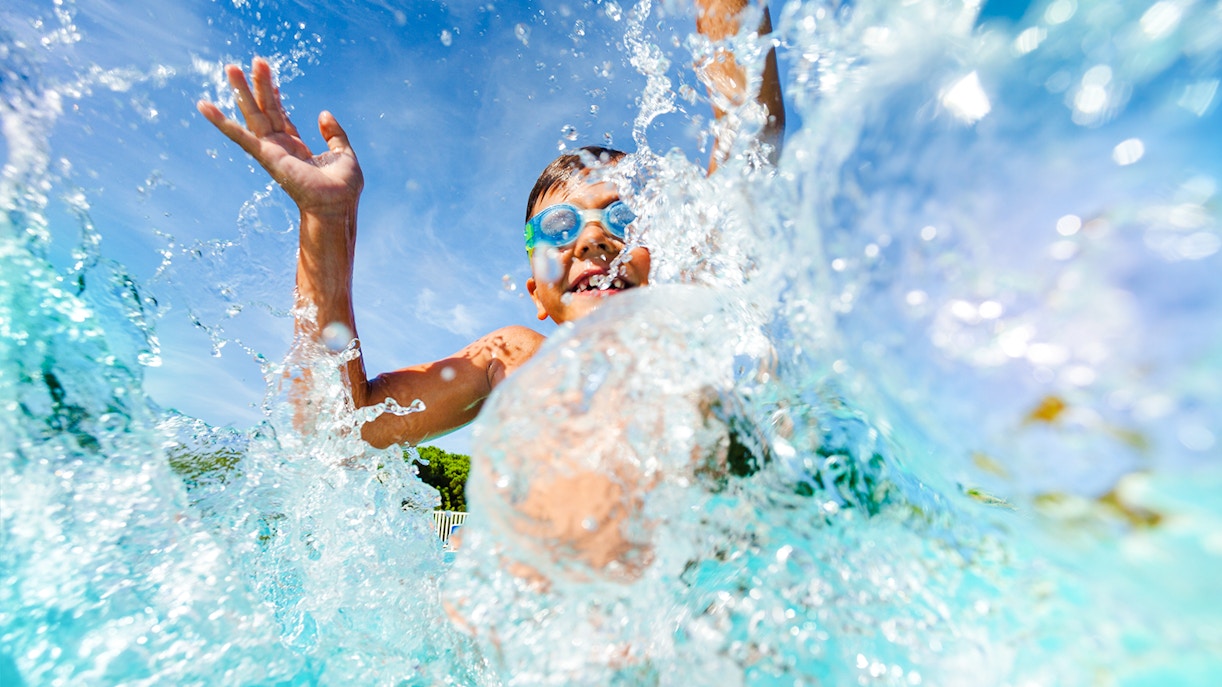 Young boy splashing in water at a water park, wearing goggles.