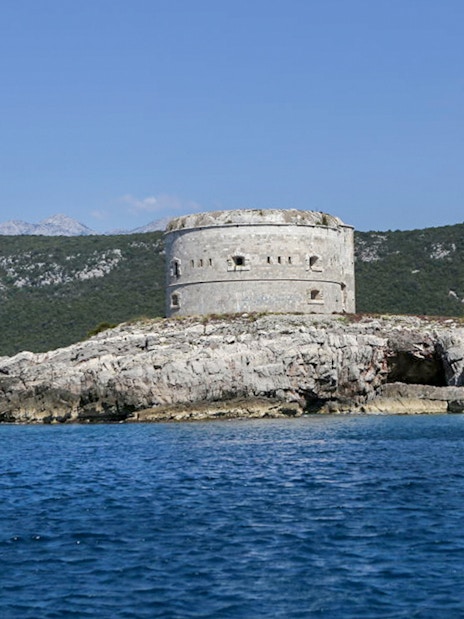 Old fort on rocky island in Boka Bay, Montenegro, with mountains in the background.