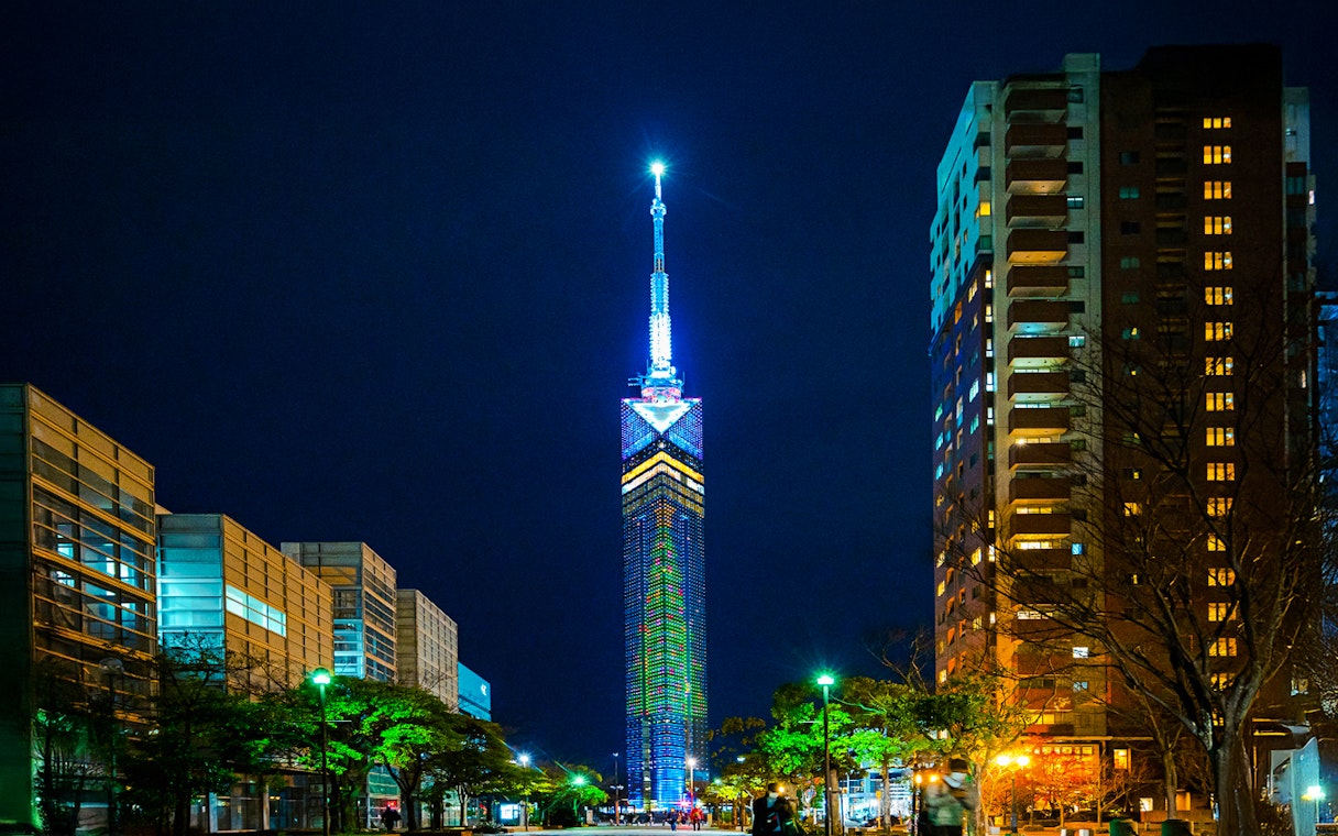 Fukuoka Tower illuminated at night, surrounded by city buildings.