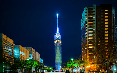 Fukuoka Tower illuminated at night, surrounded by city buildings.