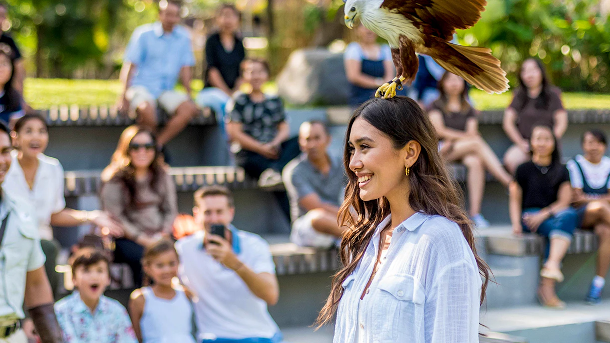 Woman smiling with a bird perched on her head at Bali Bird Park, audience in background.