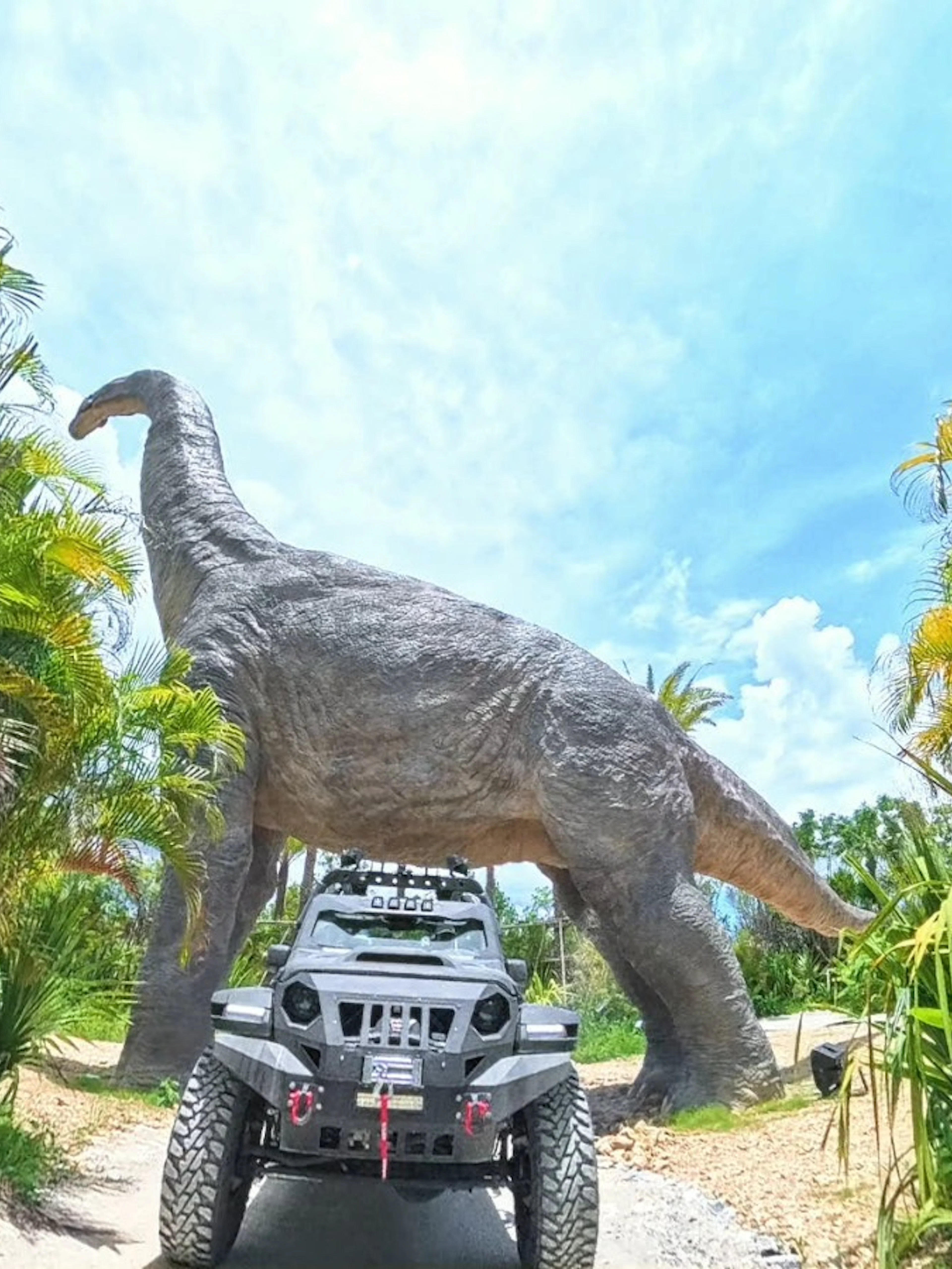 Off-road vehicle under dinosaur statue at JUNGLIA OKINAWA Park.