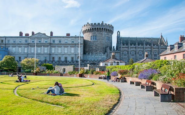 Dublin Castle gardens with people relaxing on the grass, historic stone tower in the background.