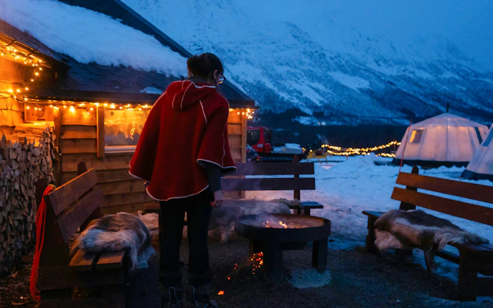 Person by a bonfire outside a cabin with snow-covered mountains in Tromso Lapland.
