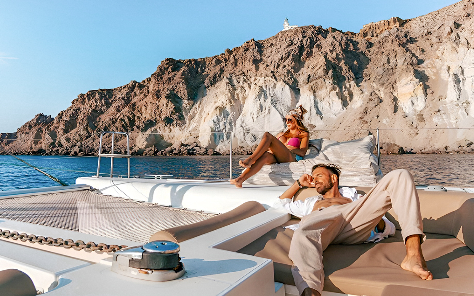 Guests relaxing on a catamaran with rocky cliffs in the background during Santorini cruise.