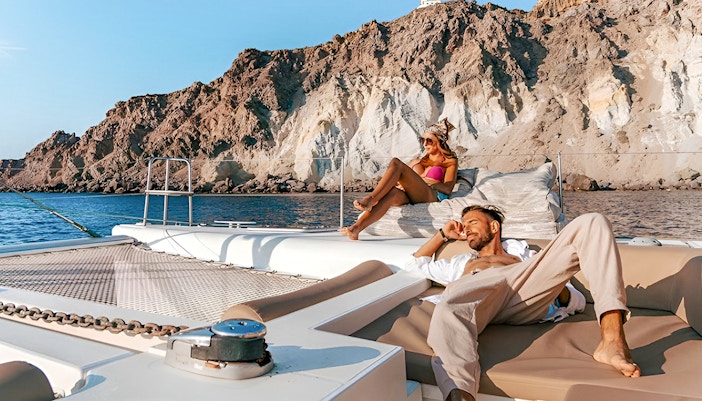 Guests relaxing on a catamaran with rocky cliffs in the background during Santorini cruise.