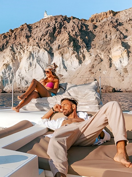 Guests relaxing on a catamaran with rocky cliffs in the background during Santorini cruise.