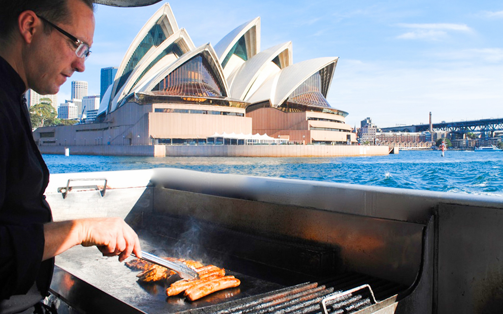 Man grilling sausages on a boat with Sydney Opera House in the background during a whale watching cruise.