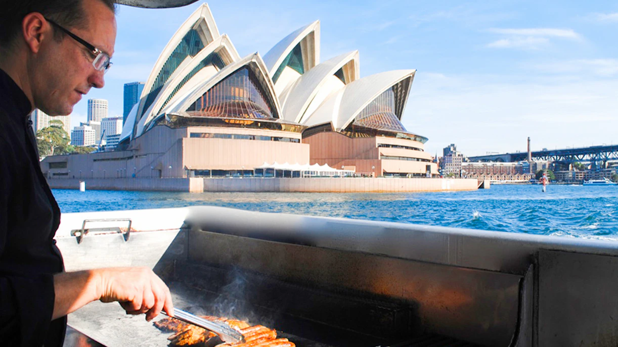 Man grilling sausages on a boat with Sydney Opera House in the background during a whale watching cruise.