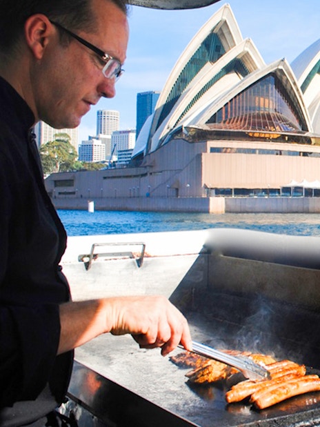 Man grilling sausages on a boat with Sydney Opera House in the background during a whale watching cruise.