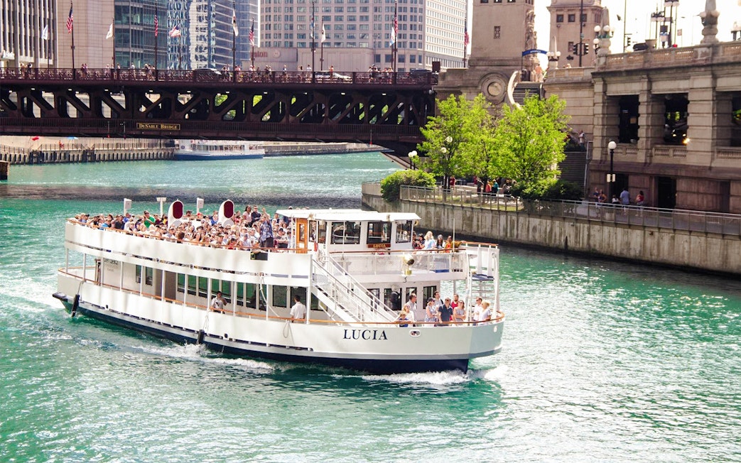 Chicago River cruise boat passing under DuSable Bridge with city buildings in the background.