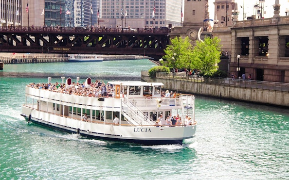 Chicago River cruise boat passing under DuSable Bridge with city buildings in the background.
