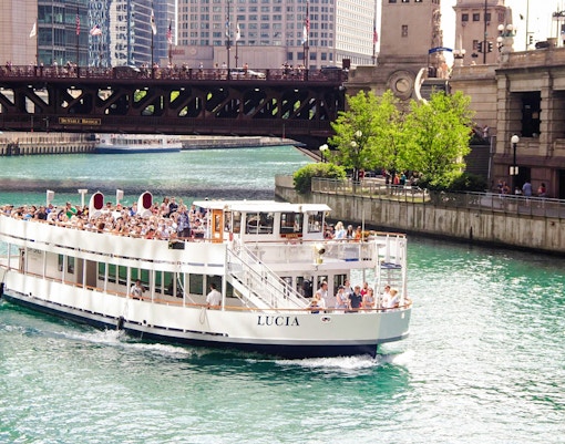 Chicago River cruise boat passing under DuSable Bridge with city buildings in the background.