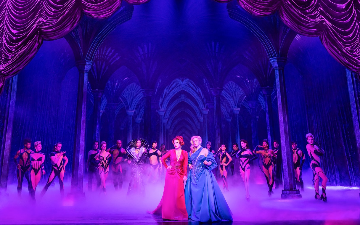 Theatrical performance of "Death Becomes Her" with actors in vibrant costumes on stage.