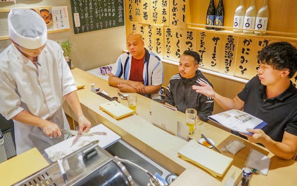 Chef preparing sushi at a counter with three guests observing in a Japanese restaurant.