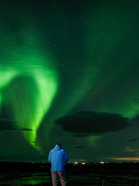 Northern Lights illuminating the night sky during an off-road tour, person in blue jacket watching.