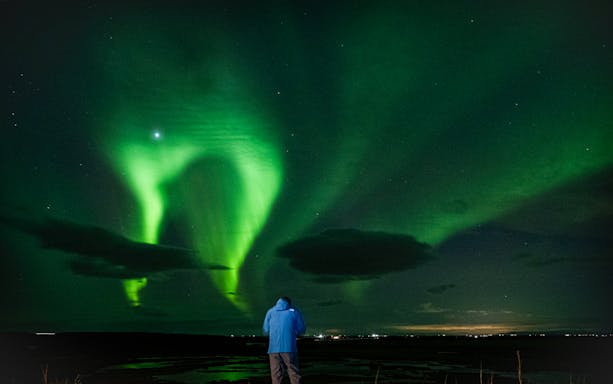 Northern Lights illuminating the night sky during an off-road tour, person in blue jacket watching.