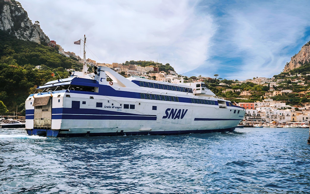 SNAV ferry approaching Capri with coastal town and cliffs in the background.