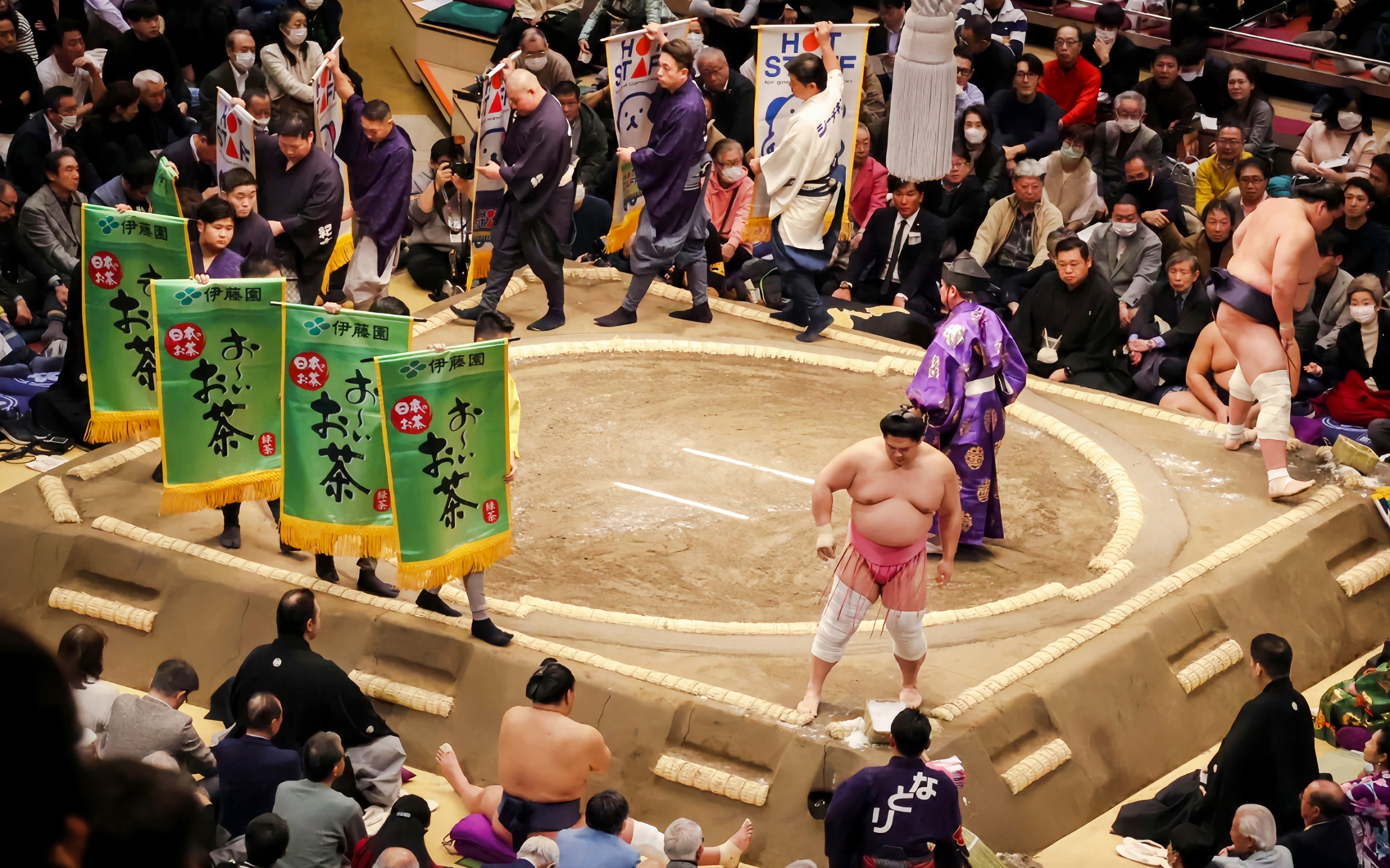 Sumo wrestler performing pre-match ritual in the ring, surrounded by banners and audience.