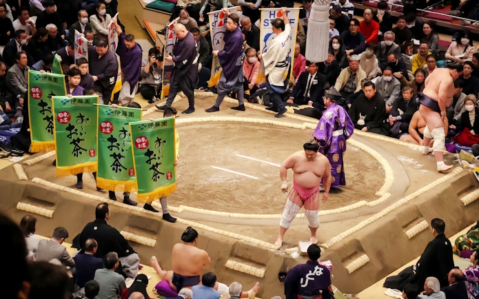 Sumo wrestler performing pre-match ritual in the ring, surrounded by banners and audience.
