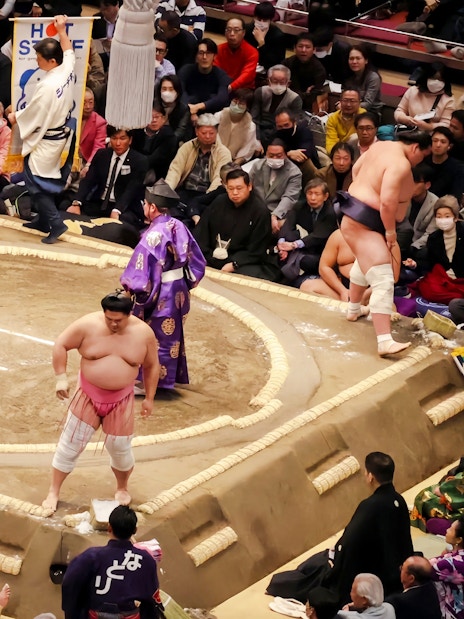 Sumo wrestler performing pre-match ritual in the ring, surrounded by banners and audience.