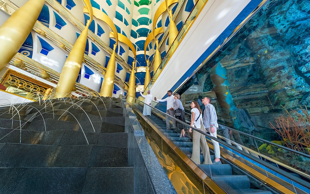 Guests on escalator inside Burj Al Arab, Dubai, with gold columns and aquarium view.